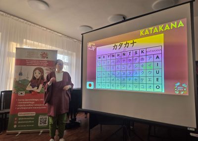 Lektorka Ola w bordowym stroju stoi przy ekranie, tłumacząc technikę pisania