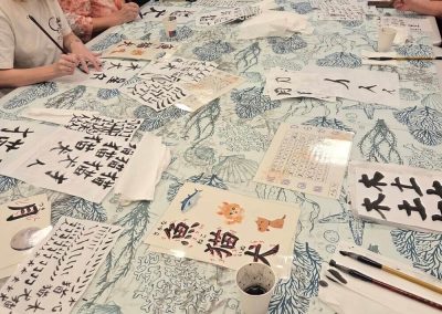 Uczestnicy piszą pędzlami na arkuszach papieru ryżowego. Na stole leżą gotowe znaki kanji (m.in. „kot”, „pies”, „ryba”), pędzle do kaligrafii, kubeczki z tuszem oraz karta z alfabetem katakana